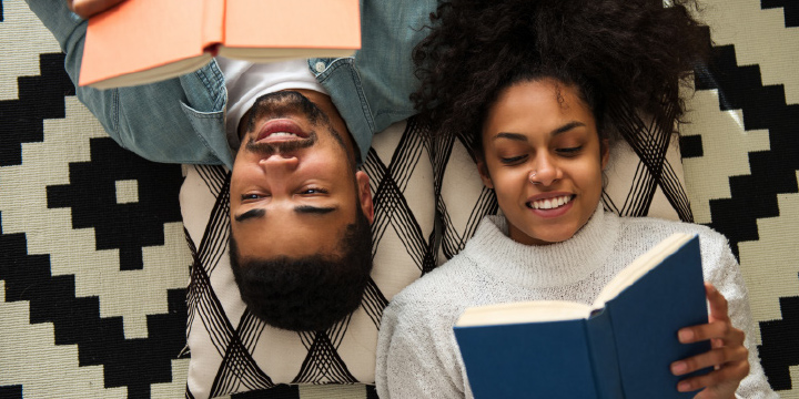 Two adults reading books while laying on a carpet, facing opposite direction.