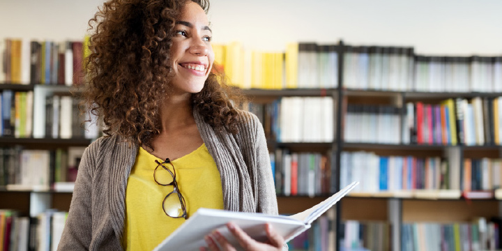 A teen girl standing in a library, holding a book, smiling.