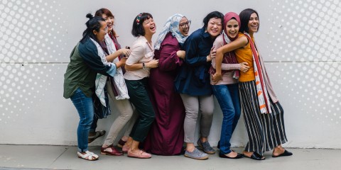 A photograph of a group of people of different cultural backgrounds standing front to back in a line laughing and smiling together
