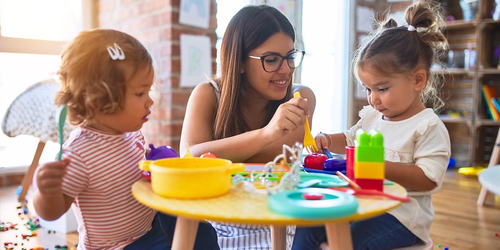 An adult sitting at a small table with two toddler. They are playing together using pretend food.