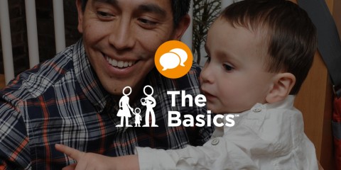 A photograph of a man holding a baby and smiling. Their bodies are facing each other, but their heads are turned together to the left and the baby is pointing at something out of frame. The logo for The Basics is superimposed on top with the icon or 