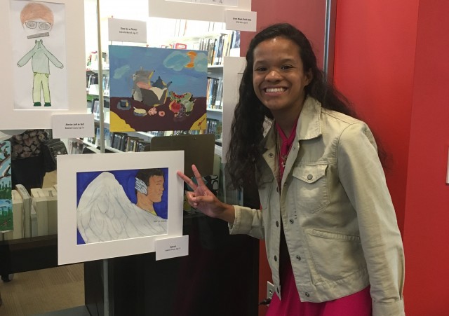 Artist displaying a peace sign next to their work, an image of a figure with large white wings.