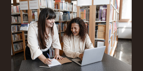 A younger and older woman work together at a table in the library. The younger woman takes notes in a notebook while the older woman works on a laptop. 
