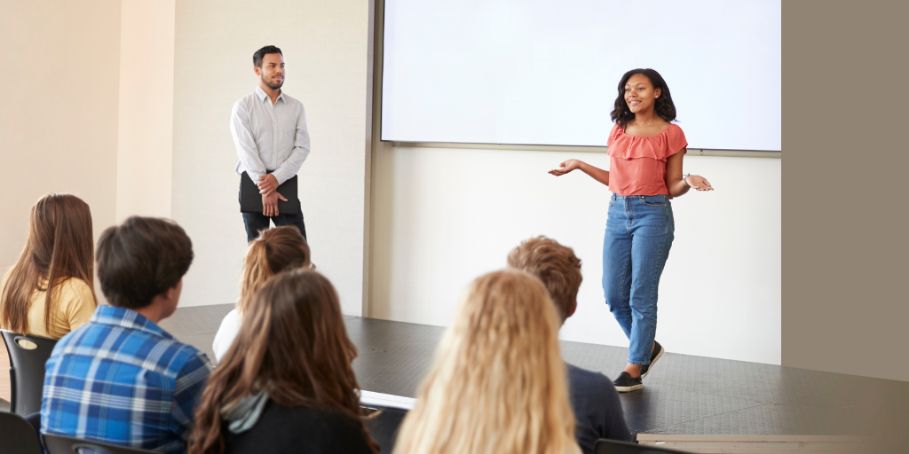 A young woman gives a presentation in front of a group of teens. The young woman stands in front of a projector screen, and a young man stands off to the side listening.  