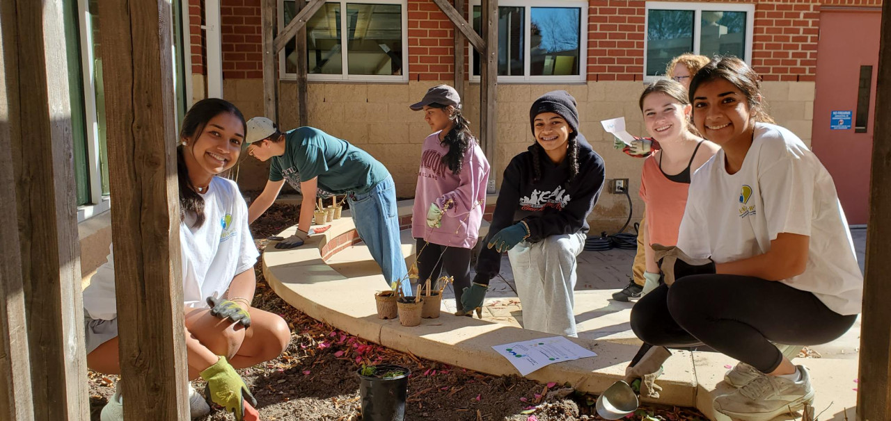 Seven teens gather around a garden bed in a library courtyard. In the foreground, four teens stop and smile at the camera while working. In the background, three teens continue digging and observing the garden.