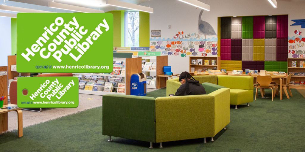 A wide angle photograph showcases the children's area of a library. The camera captures the back of a child who is sitting on a green couch. Beyond the child are rows of book shelves that are low for children to browse. The wall features several colorful cut out fish and a blue heron.   