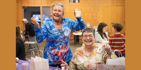Two older adult women smile at the camera while holding up paint brushes and small gardening pots with painted designs. The woman standing on the left wears a colorful printed shirt with sea creatures, and the woman sitting on the right wears a shirt with a colorful floral pattern.