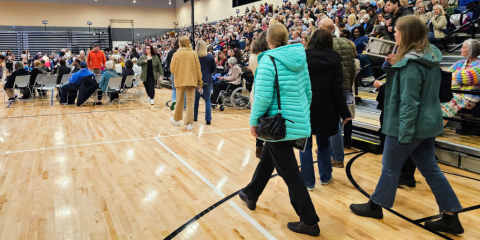 In the foreground, a group of individuals walks into an arena with a crowd of people filling floor seating and bleachers.