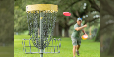 Image foreground shows a close-up of a disc golf basket. In the background, blurred, is an adult throwing a disc golf disc aimed toward the basket. Behind the adult are several large trees and other greenery.