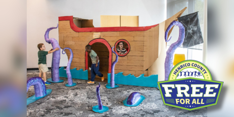 Three children play in and around a large cardboard pirate ship in a library meeting room. Scattered around the pirate ship are several purple papier mache tentacles emerging from the water/floor.
