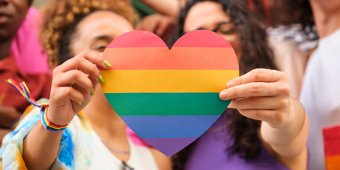 The foreground features a paper rainbow heart in sharp focus. In shallow focus, behind the heart, are the two women holding it. Each woman holds one side of the heart.