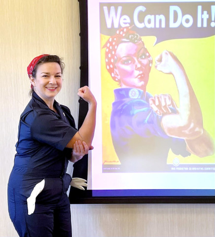 A presenter dressed as Rosie the Riveter stands in front of a projector displaying the famous Rosie the Riveter artwork with the slogan, 