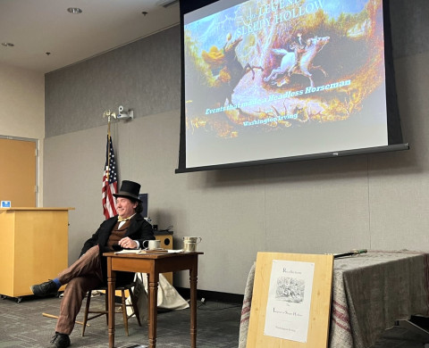 A presenter dressed as Ichabod Crane sits at the front of a meeting room in front of a projector. Text on the projector reads, 