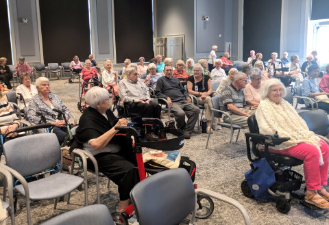 Several members of a senior living community sit in a large meeting space for a presentation. 
