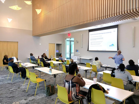 A presenter stands in front of a large library meeting room. Behind her is a projector screen with information about the Henrico Economic Development Authority. The audience sits at three rows of tables set up in front of her.