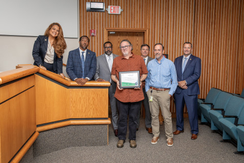 Seven individuals stand at the front of a boardroom and pose with an award.
