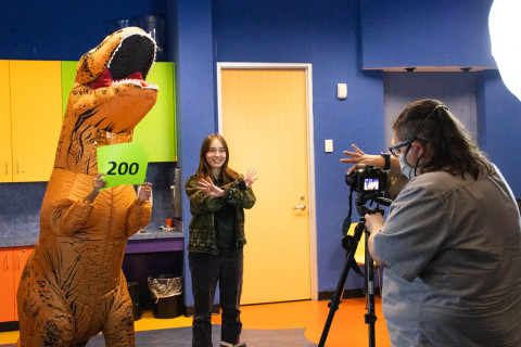 In the foreground a librarian adjusts a video camera on a tripod. In front of the camera is a teen smiling with her arms criss-crossed. She stands next to a teen in an inflatable dinosaur suit holding a lime green sign with 