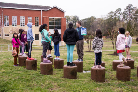Teens and tweens stand on concreate stools outside of Tuckahoe Library while looking and listening to staff talk about community science.