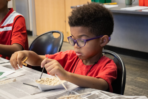 A young boy wearing glasses sits at a table and uses a brush and spoon to dig through a container for various dinosaur trinkets. 