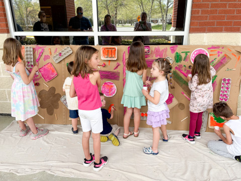 Eight young children stand and kneel in front of a large piece of cardboard, where they paint, draw, and add objects to the mural project. 