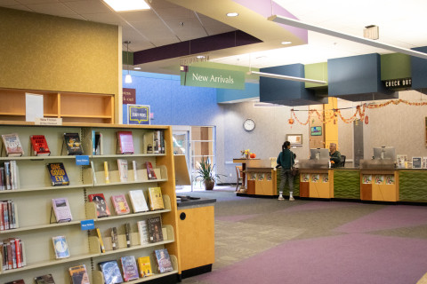 A wide-angle photograph shows bestseller book shelves in the foreground and the Circulation desk with a patron and staff member in the background.