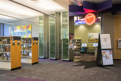 A wide-angle photograph features the entrance to the Teen area of the library. Above the entrance is a blue and red neon light sign that reads 