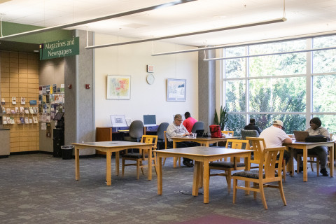 A wide-angle photograph shows several patrons seated at tables reading and working in the Magazine and Newspaper area of the library. Beyond them are large windows with a view overlooking the garden behind the library.
