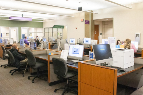 A wide-angle photograph features rows of public use computer at the library. Patrons are seated at a few of the stations. In the background a sign reads, 