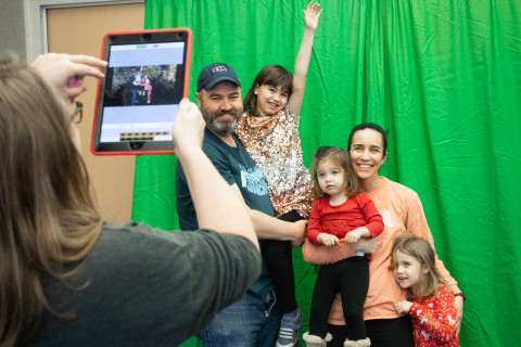 Two adults and their three young children pose in front of a green screen as a staff member takes their photo with an ipad. A background of fireworks replaces the green screen.