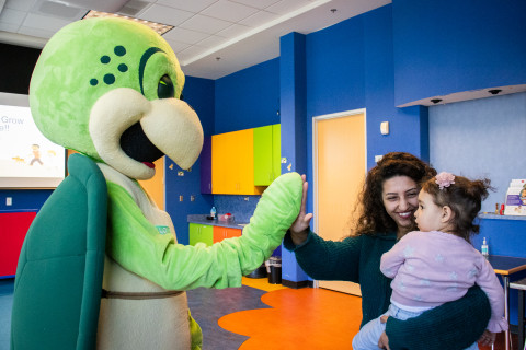 A woman carrying a young child gives a high five to a turtle mascot. Both the turtle and the woman smile at the young child, who looks at them with a curious expression.