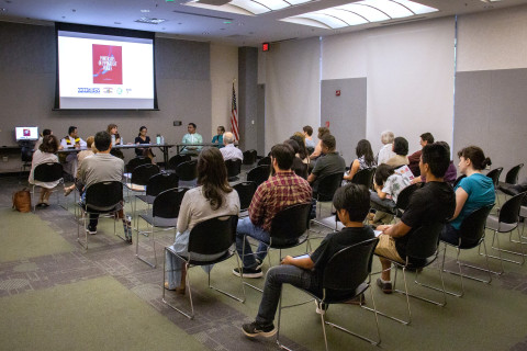 An audience of approximately two dozen patrons sits and listens to a panel of library staff and community members discussing the immigrant experience. Behind panel members is a projector with a slide featuring the cover of the book, 