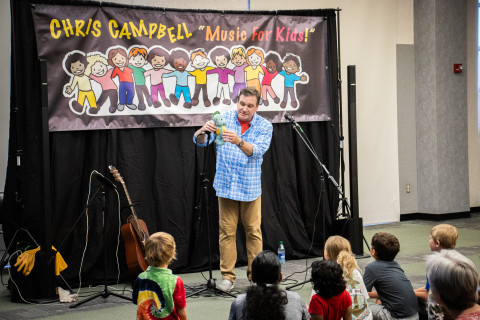 Musical entertainer Chris Campbell stands in front of a crowd of children holding a stuffed animal. Beside him is his guitar.