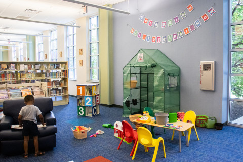 A wide-angle photograph of a young child playing in the Children's area of Tuckahoe Area Library. Toys are scattered across the floor and across a children's table.