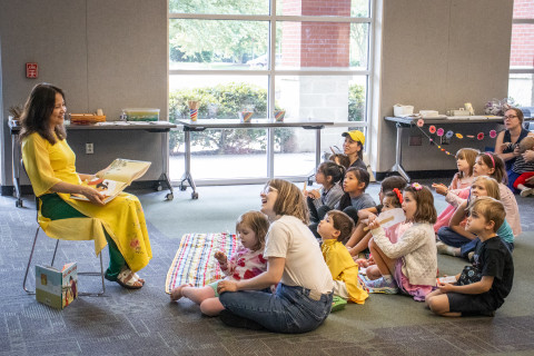 A group of children and their caregivers sit on the floor. They watch and listen to author Hanh Bui read one of her books.