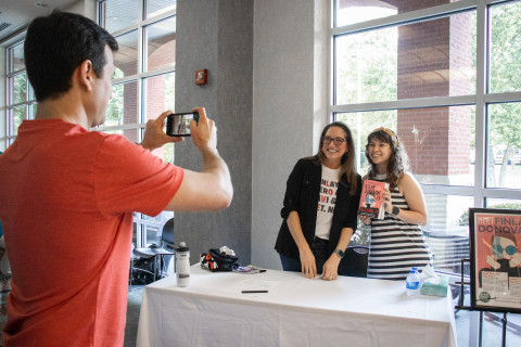 A patron poses for a photo with author Elle Cosimano. They stand behind a book signing table while another patron takes the photo for them.