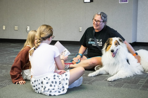 Two teens sit on the floor and read aloud to a large white and brown collie.