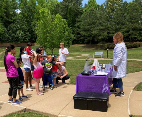 A group of teens and tweens stands outside. They view a presenter who is wearing a lab coat and conducting an experiment on the table in front of them. 