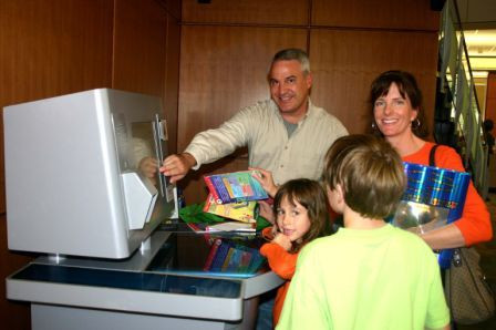 A family smiles at the camera while using the library's self checkout machine.