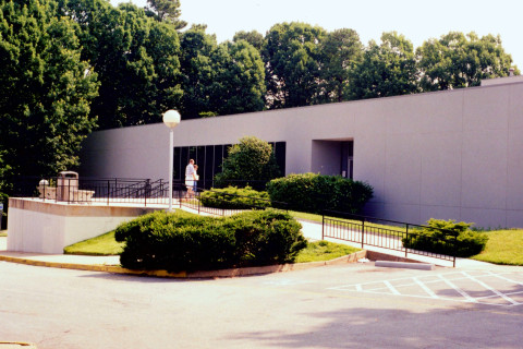 An exterior view of a gray office building with a ramp leading up to its entrance. At the top of the ramp are two benches and a trash can. The photo captures a patron walking toward the building.