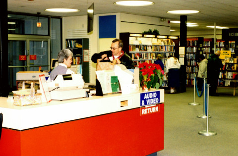 A staff member assists a patron at a library reference desk. A sign on the desk reads, 