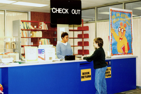 A staff member assists a child at a library reference desk. A sign above the desk reads, 
