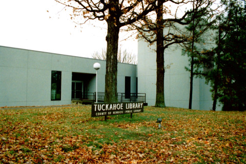An exterior view of a gray office building. In front of the building are a few tall pine trees and a sign that reads, 