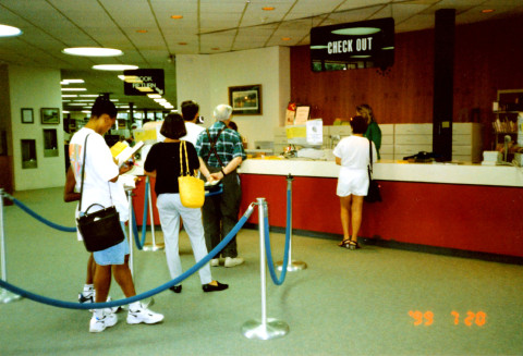 A line of people waits behind rope stanchions at a library circulation desk. A staff member assists a patron at the desk.