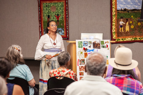 Artist Denise Lowe Walters stands in front of an audience. Behind her are her story quilts. Next to her is a bulletin board with different samples of her quilting materials.