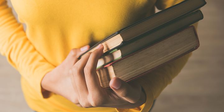 A photo of a woman in a yellow shirt holding three books in her arms.